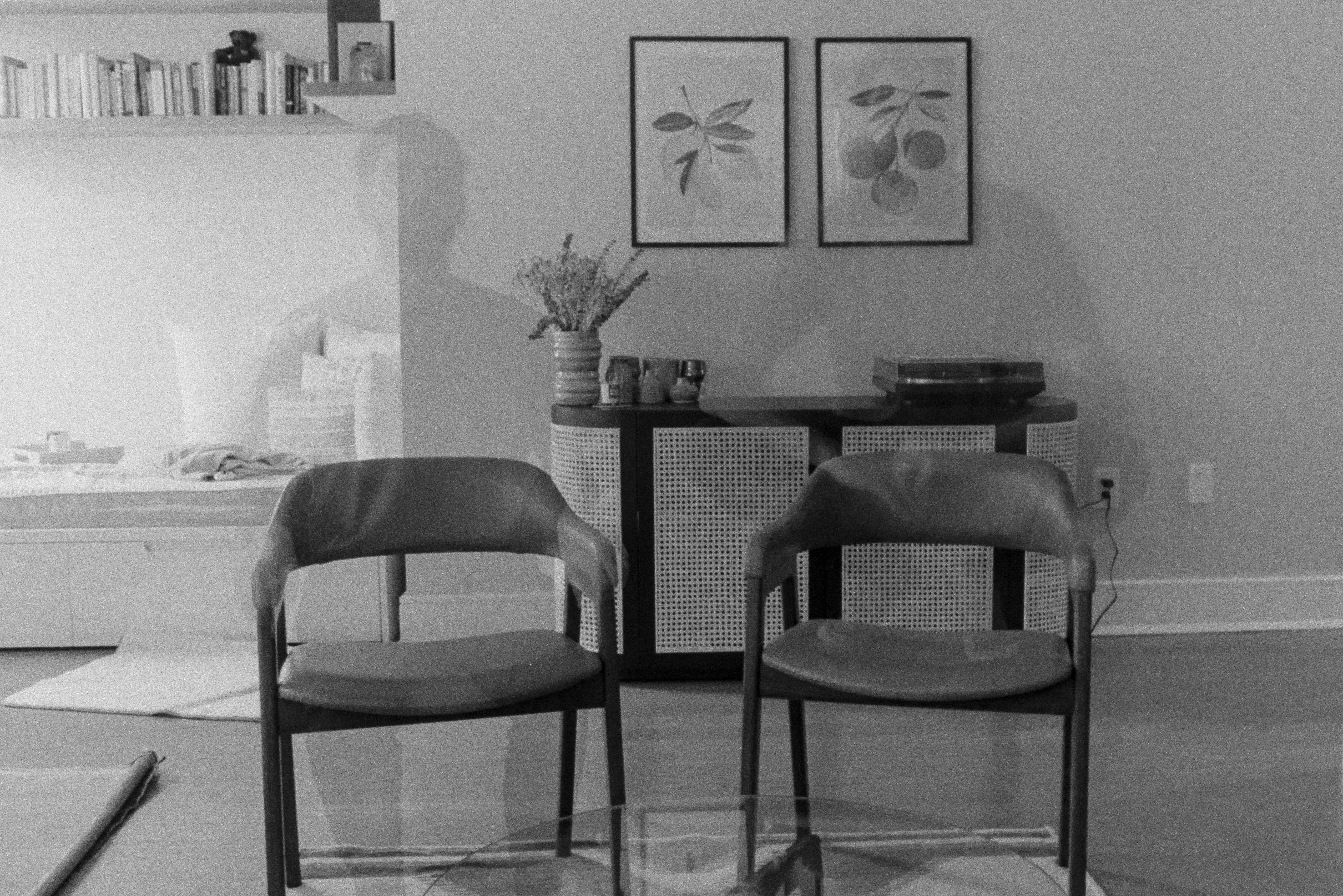 A black and white self portrait. Ghost-like image of a man sitting in a chair and the same man squatting on the chair next to him.